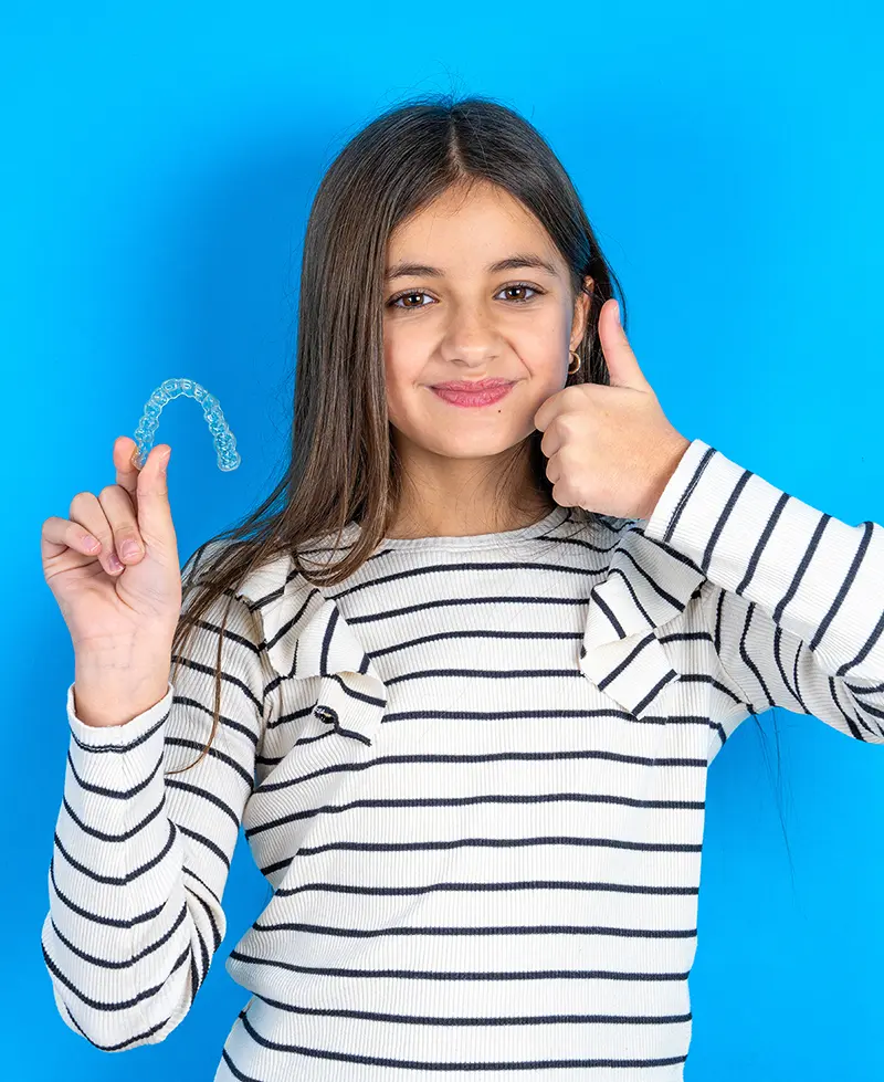 a girl holding a clear retainer, smiling and doing a thumbs up against a blue background
